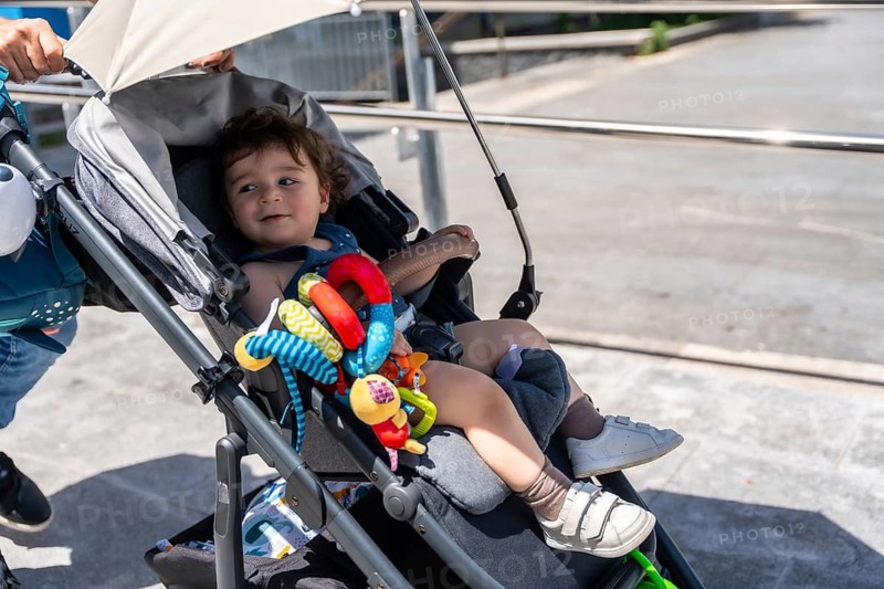 Curious baby boy sitting in a stroller with a colorful toy, smiling and looking away during a sunny outdoor walk in the city park, enjoying a relaxed family outing