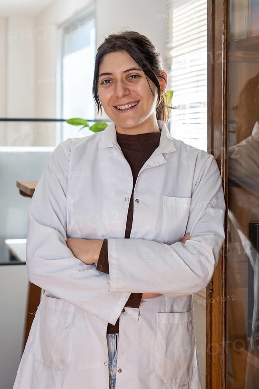Young female scientist in white lab coat smiling broadly with arms crossed, leaning against a wooden cabinet in a bright laboratory office with indoor plants