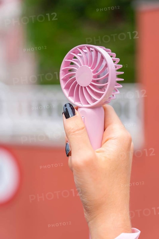 Person's hand gripping a pink portable mini fan, providing personal cooling and relief during hot outdoor weather, symbolizing comfort, travel, and modern electronics