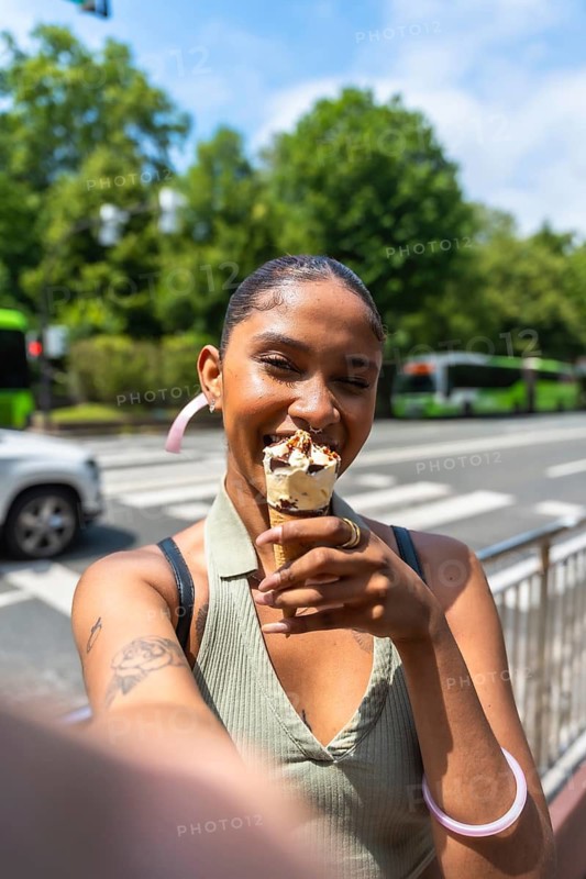 Happy young woman smiling with an ice cream cone, capturing a selfie on a vibrant city street during a sunny summer day, embodying joy and urban lifestyle