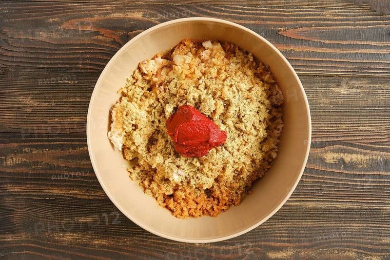 Bowl on wooden table filled with minced meat, boiled rice and spices. Red tomato sauce placed in center. Preparation for cooking takes place in kitchen