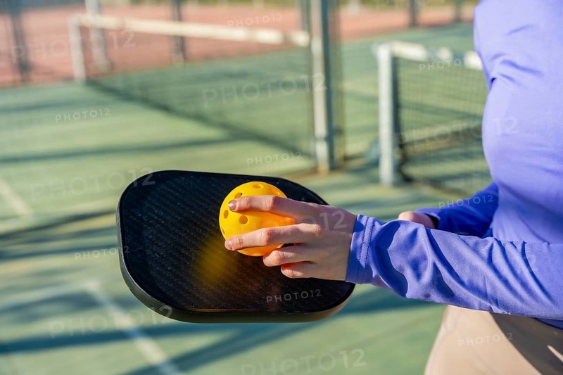 Person is holding a black pickleball paddle and a yellow pickleball ball, preparing to play on an outdoor court, symbolizing leisure, healthy lifestyle, and athletic activity