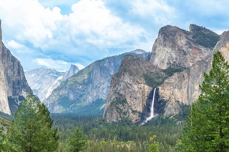 Yosemite valley panorama presenting towering granite cliffs like el capitan and half dome, with bridalveil fall cascading into a lush evergreen forest under a bright sky with light clouds