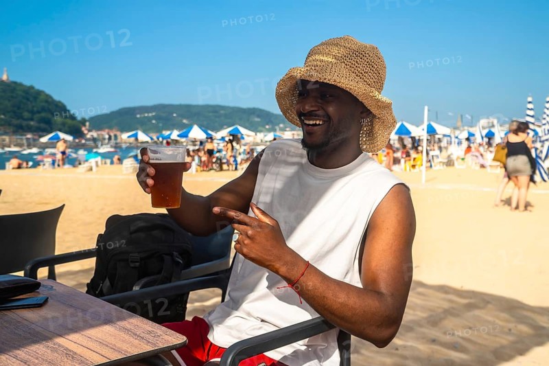 Happy young man wearing a straw hat and drinking a fresh beer at a beachside bar, relaxing while pointing at something with the sandy beach and clear blue sky in the background
