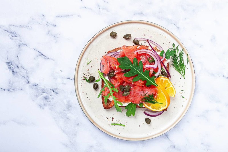 Sandwich with freshly smoked salmon, on black bread, with cottage cheese, with herbs and red onions, capers and pieces of citrus fruits, on a marble countertop, breakfast, no people