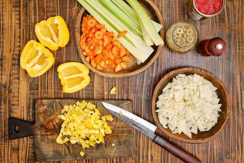 Vegetables are prepared on a wooden table. Yellow pepper slices are next to chopped onions and diced yellow bell peppers. Celery sticks and carrot pieces are also arranged in bowls