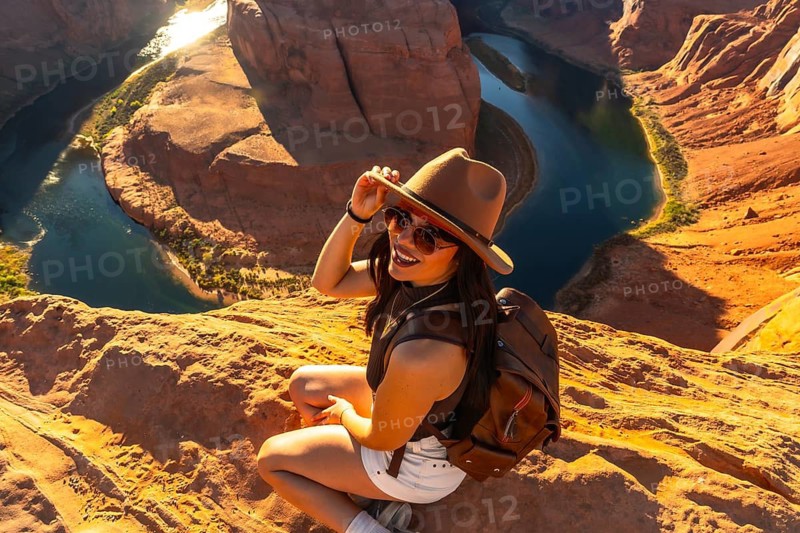 Woman in hat and sunglasses sits on sandstone cliff edge smiling at camera, overlooking the panoramic horseshoe bend and the colorado river winding through red rock canyon in arizona