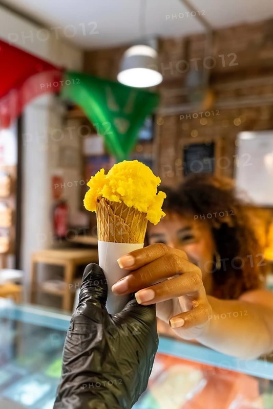 Shop worker wearing a glove passing a scoop of refreshingly sweet yellow fruit ice cream in a crisp waffle cone to a happy customer at the counter, symbolizing summer treats and service