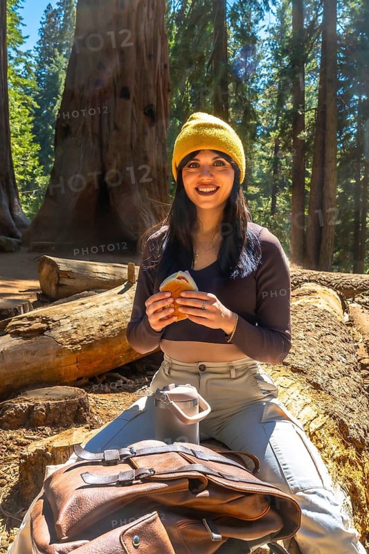 Young woman smiling while sitting on a large fallen sequoia log, eating a sandwich and a drink in a thermos, taking a break during her hiking trip in a giant sequoia forest