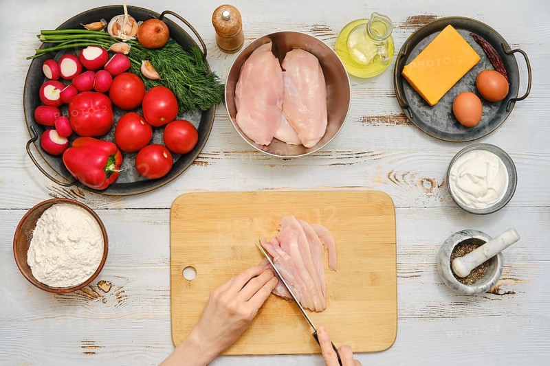 A person is cutting chicken on a wooden board. Various fresh vegetables like tomatoes, radishes, and peppers are nearby. Bowls hold eggs, cheese, and other cooking items in a kitchen space