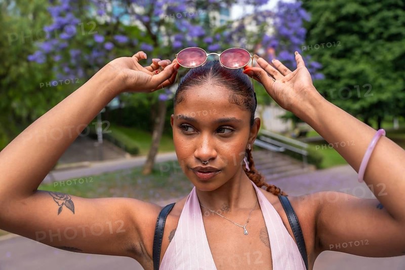 Natural young woman with dark skin and braided hair raising pink sunglasses on her head, posing outdoors on a sunny day with blooming jacaranda trees