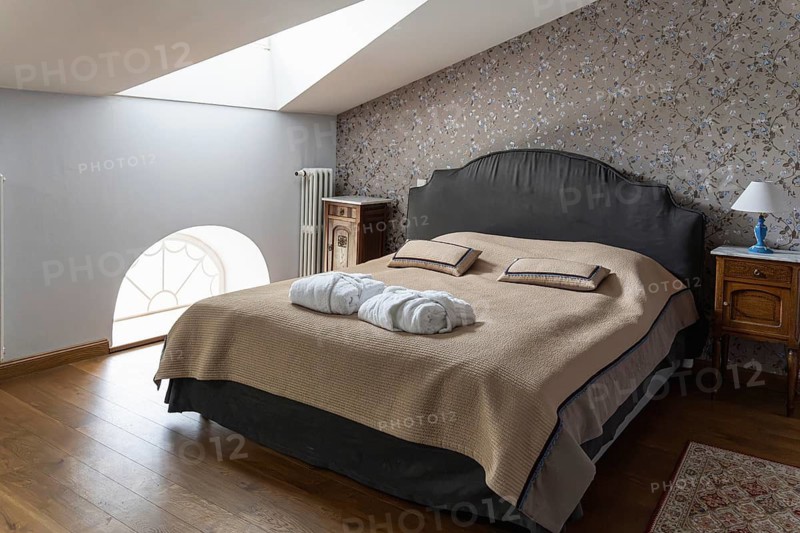A cozy attic bedroom features a dark upholstered bed with a beige bedspread and matching pillows. Two folded white bathrobes lie neatly on top. Wooden nightstands stand on each side, one with a blue lamp and the other near a decorative cabinet and radiator. A skylight brings natural light into the room, highlighting floral wallpaper, wooden flooring and a patterned rug. The space blends comfort with classic decorative elements