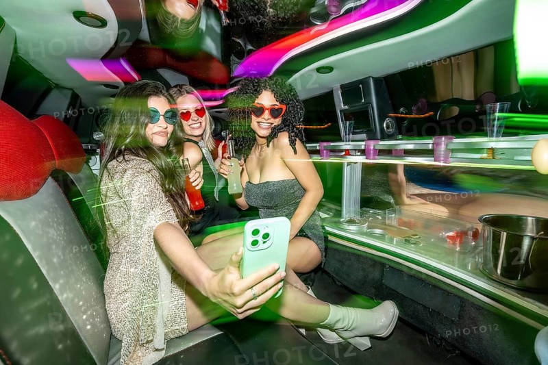 Three diverse young women wearing party dresses and heart shaped sunglasses, enjoying a luxurious limousine ride with drinks and taking a selfie, capturing moments of fun and celebration
