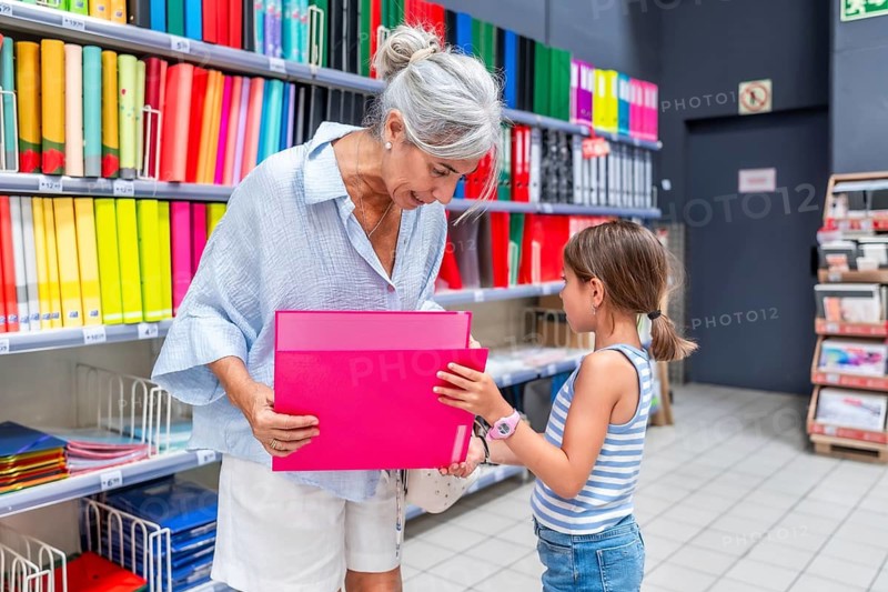 Grandmother and granddaughter shopping for school supplies together, with the older woman holding two pink folders while the young girl points at items on the colorful shelves
