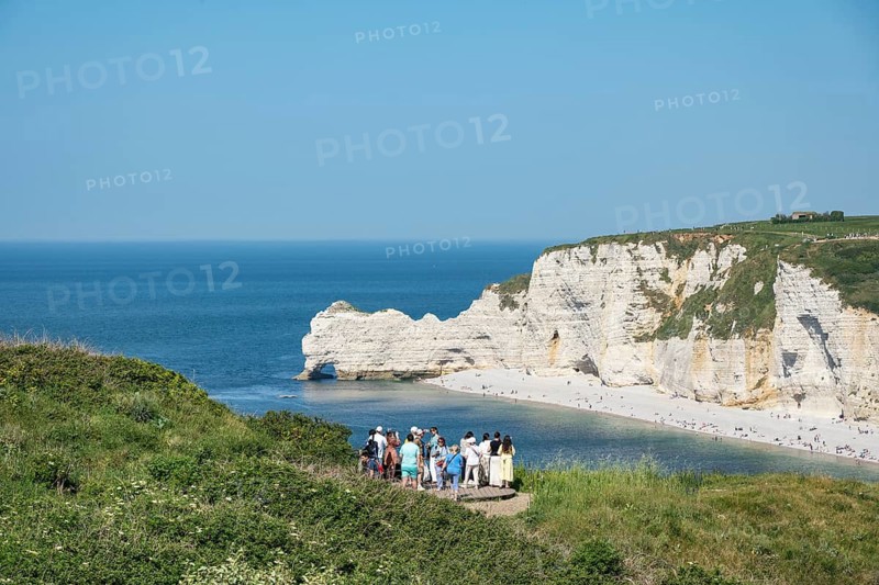 Tourists stand at a viewpoint on the Falaise d'Aval and look at the pebble beach of the seaside resort of Étretat and the overlying rock formation and landmark Falaise d'Amont on the English Channel, Alabaster Coast, La Côte d'Albâtre, Chalk Cliffs, English Channel, Seine-Maritime, Normandy, France