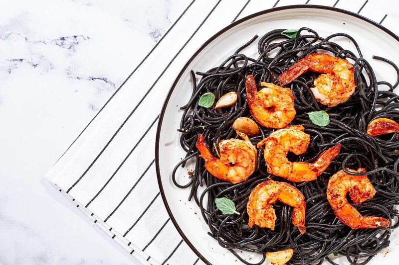 Black spaghetti with shrimp, on a white plate with a fork, on a striped napkin, marble background, homemade, no people