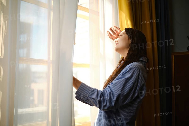 Woman stands by window, pushing back white curtain with one hand. Sunlight enters room, illuminating her face. She seems to enjoy time spent in this bright moment, relaxing in cozy space