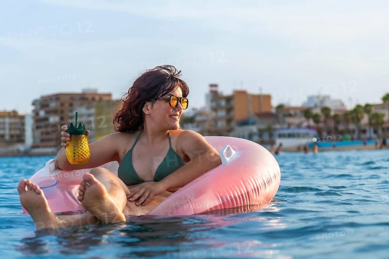 Woman smiling and enjoying summer vacation, floating in the sea on a pink inflatable ring while holding a refreshing drink in a pineapple shaped cup