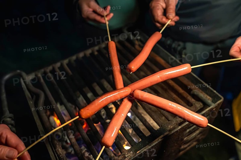People holding skewers with hot dogs over a glowing campfire grill grate, cooking and enjoying a casual outdoor meal together during a summer evening camping gathering
