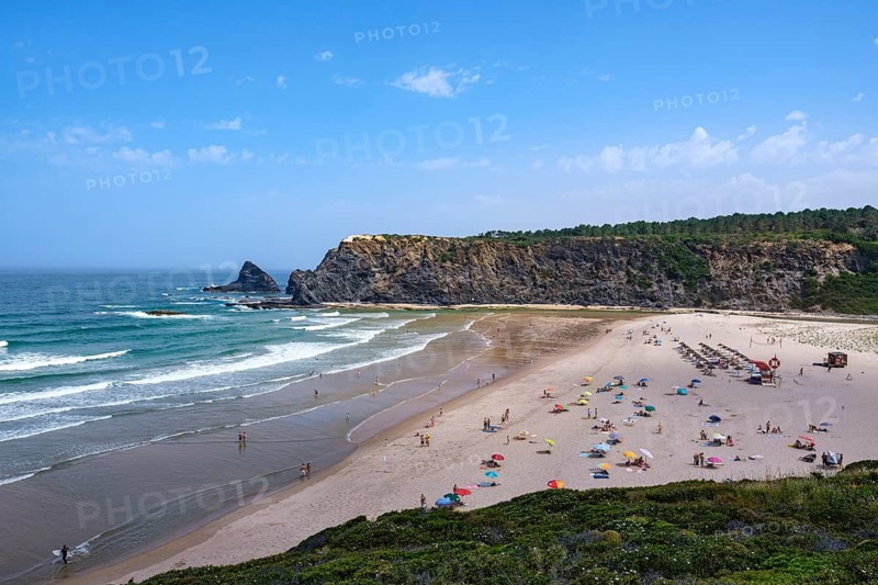 A busy beach with colorful umbrellas and rock cliffs under a blue sky, Odeceixe, Faro, Portugal
