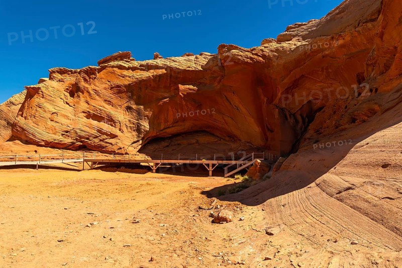 Biidi arch cave in page, arizona, displaying a striking sand colored canyon landscape, with a wooden boardwalk leading into the natural rock formation against a clear blue sky