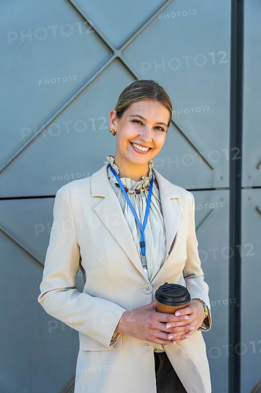 Young businesswoman smiling, holding a takeaway coffee cup, wearing a smart blazer and lanyard, standing confidently outdoors against a modern gray wall