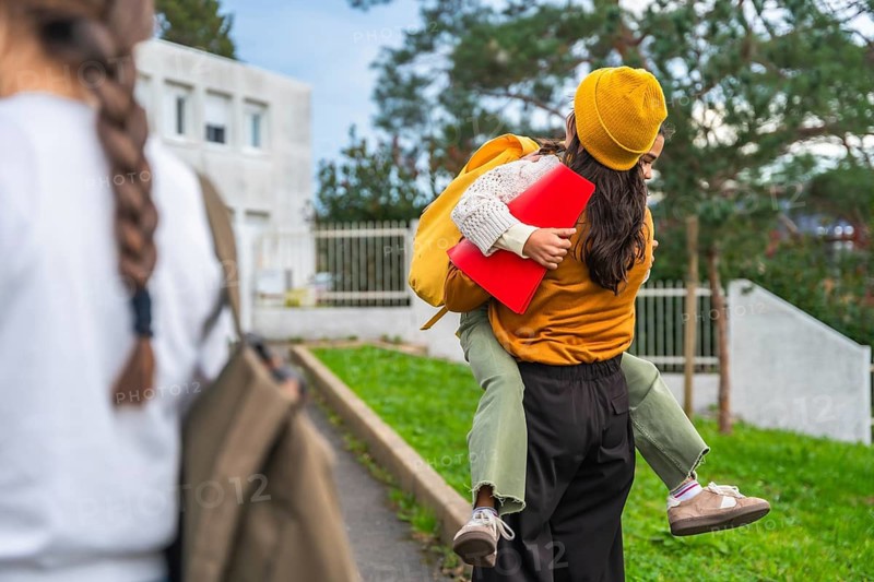 Mother carries her smiling daughter on her back after school, sharing a joyful piggyback hug as they walk home through a neighborhood, showing love and everyday bonding