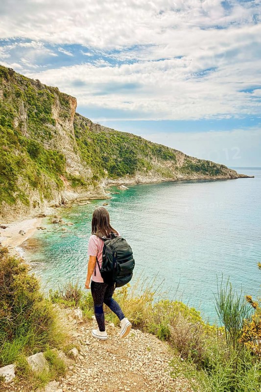 Aerial view woman hiking on viewpoint isolated go down to Filikuri beach in spring. Clear turquoise Ionian sea meets steep rocky coastal cliffs. Scenic Albanian Riviera travel landscape in Himar
