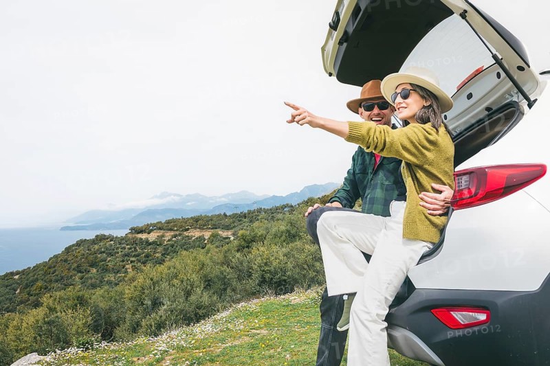 Happy couple sits in their white rented car trunk, laughing, enjoy nature views during spring road trip. Lifestyle shot capturing joy, friendship, and outdoor adventure with scenic hilly landscape