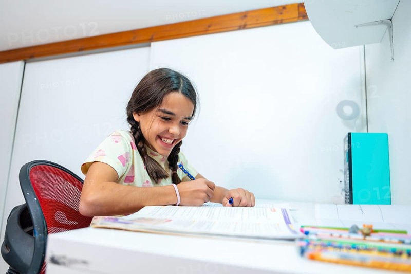 Happy schoolgirl with braided pigtails smiling as she studies and writes in a notebook at her white desk, focused on homework in a bright home learning space