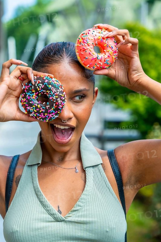 Young woman playfully covering her eyes with two vibrant sprinkle covered donuts while expressing joy and fun outdoors, embracing a sweet and cheerful moment