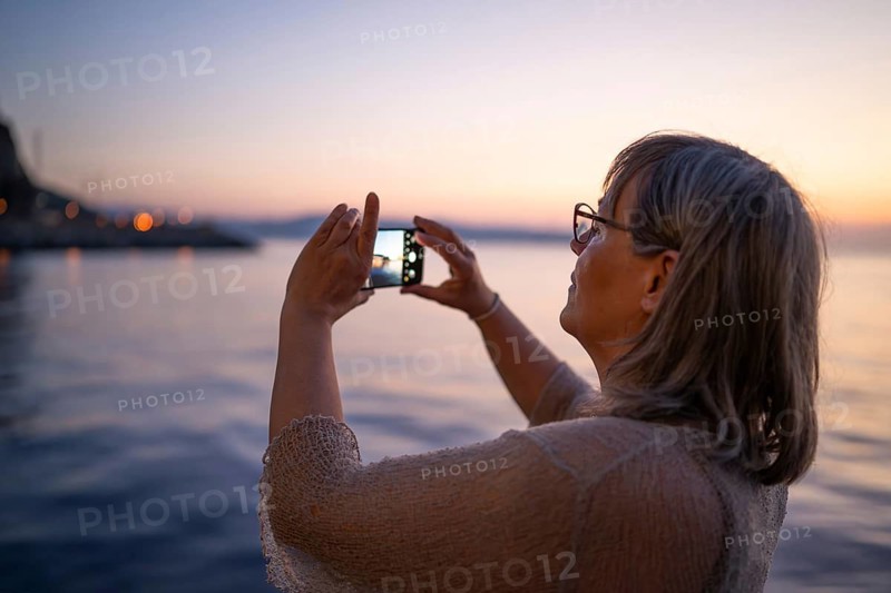 Woman with cell phone taking pictures of coast at sunset, Strait of Messina, Calabria, Aeolian or Aeolian Islands, Sicily, southern Italy, Italy