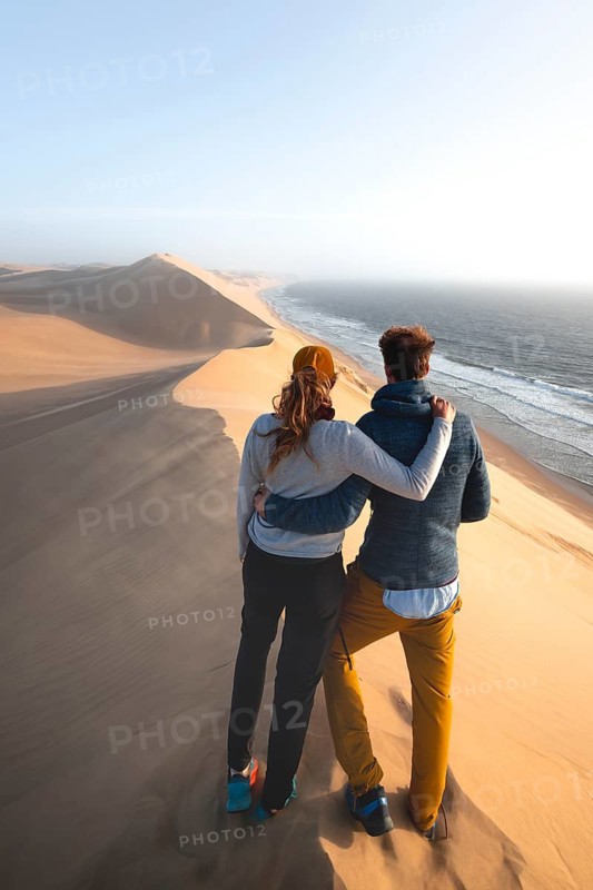 Young couple standing on a dune in front of an impressive desert landscape, sand dunes and sea in the evening light, wind blows sand across the dunes, Atlantic coast and sandy desert with sea of dunes, Sandwich Harbour, Namib Naukluft Park, Namib Desert, Namibia