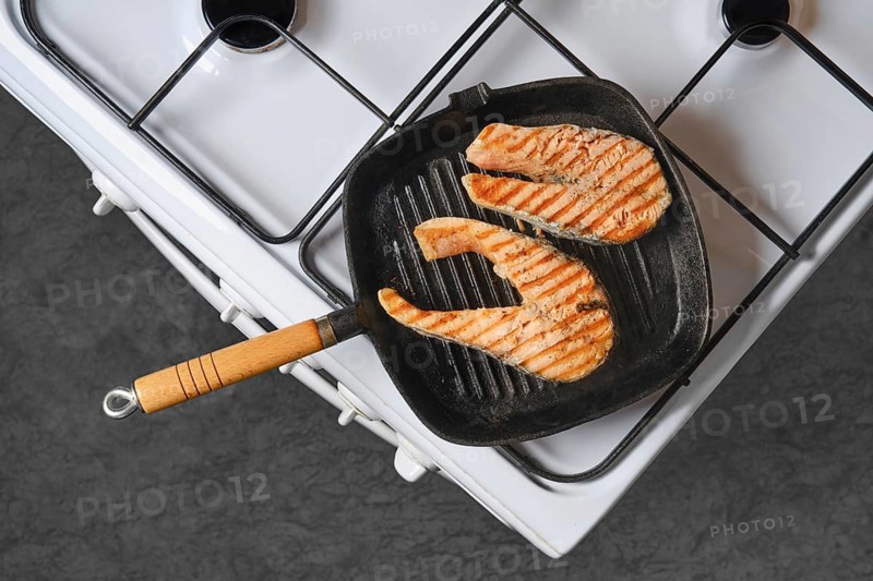 Salmon fillets are cooking on grill pan placed on stovetop. Grill marks are visible on fish. Kitchen background shows stove and counter. Cooking time is midday