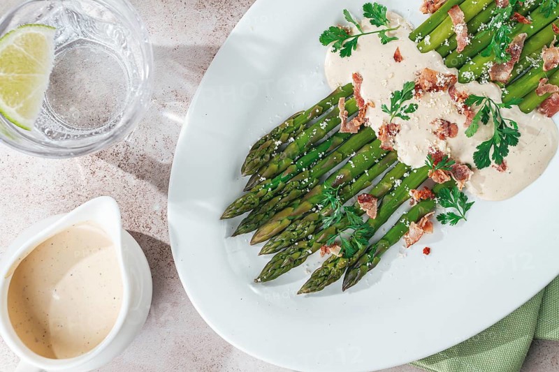 Fresh asparagus with cream sauce and bacon, on a white oval plate, light marble table, sunlight, homemade, no people