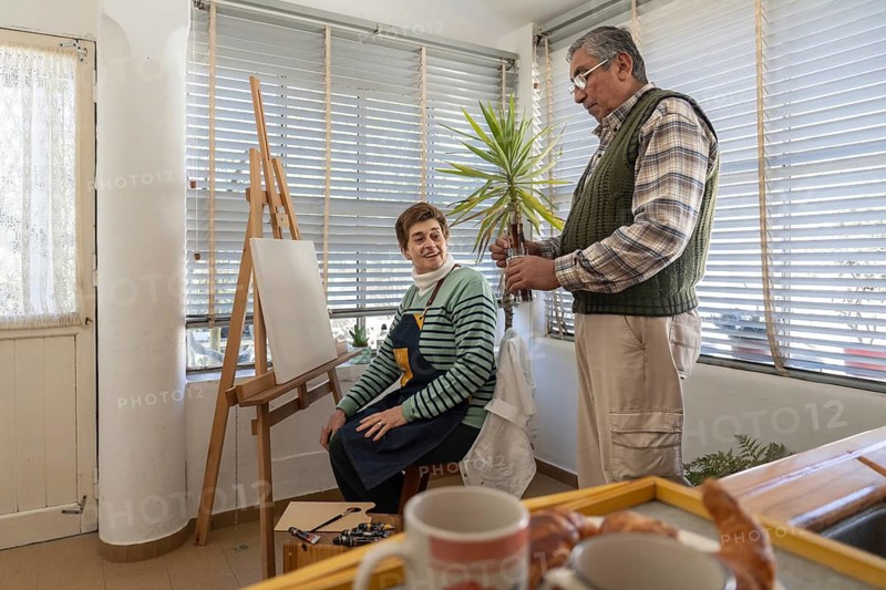 Wide shot of a senior woman seated at her easel laughing with her standing husband in a bright home studio, with a wooden breakfast tray holding coffee mugs and a croissant in the foreground