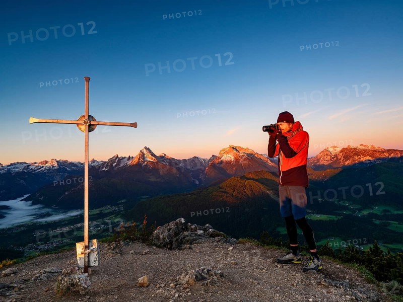 Photographer at Rauher Kopf summit cross at sunrise, in the background the Berchtesgaden Alps, Steinernes Meer, Schönfeldspitze, Watzmann, Hochkalter and horse-rider Alpe, Berchtesgaden National Park, Bischofswiesen, Berchtesgadener Land, Upper Bavaria, Bavaria, Germany