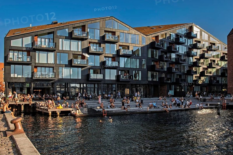 Menschen entspannen am Wasser vor einer modernen Glasfassade in der Abenddämmerung, Kopenhagen