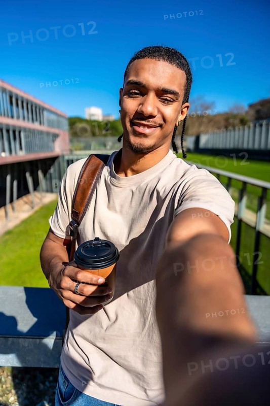 Young male student with dreadlocks and backpack, holding a reusable coffee cup and taking a selfie on a sunny university campus between classes, smiling and confident