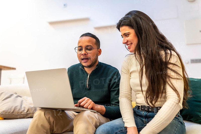 Young man and woman sitting on a sofa, collaborating on a project and smiling while looking at a laptop computer screen at home, representing teamwork and remote work