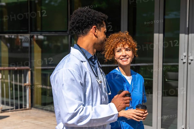 Medical team members, a male doctor wearing a white coat and a female nurse in blue scrubs, smiling and communicating while holding takeaway coffee cups during a break outside the hospital