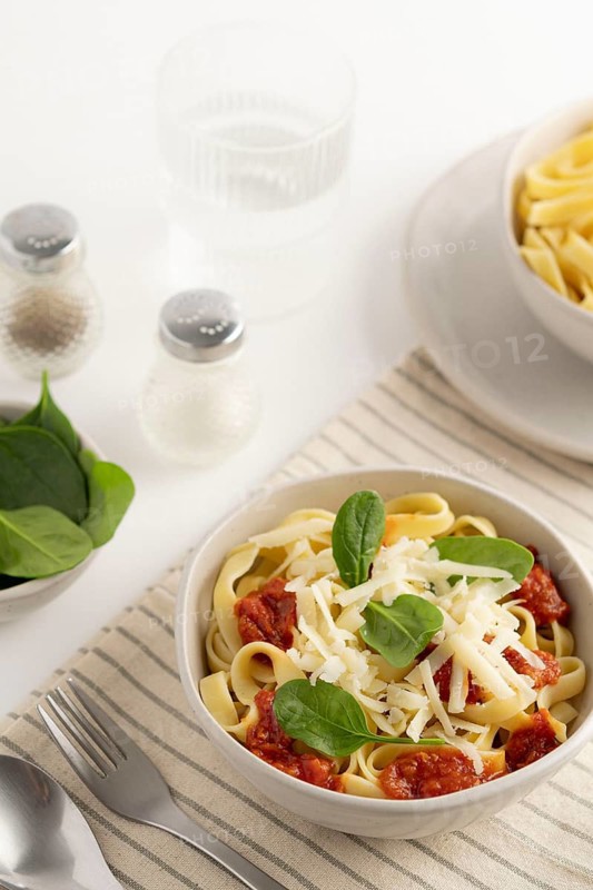 A bowl of spaghetti topped with tomato sauce, grated cheese, and fresh spinach leaves. Beside it, a glass of water, salt, and pepper shakers on a striped cloth