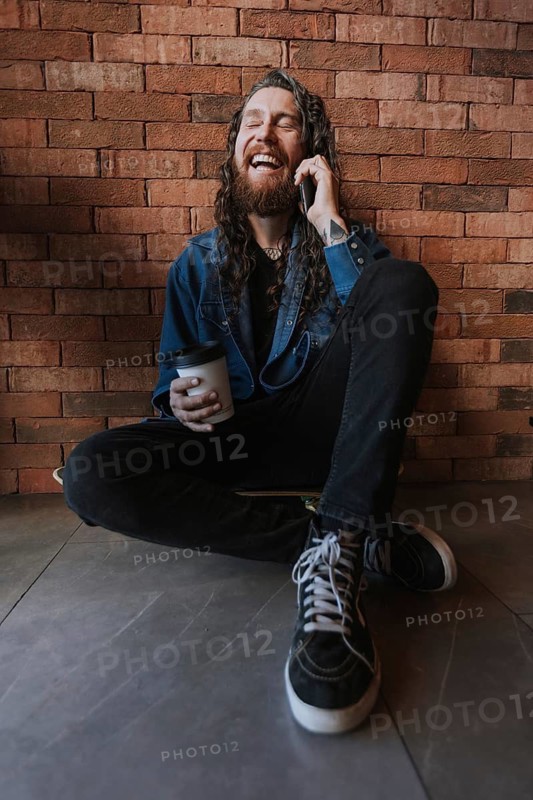 A man with long hair and a beard laughs heartily, sitting against a brick wall. He holds a phone to his ear and a cup of coffee, wearing casual jeans and sneakers