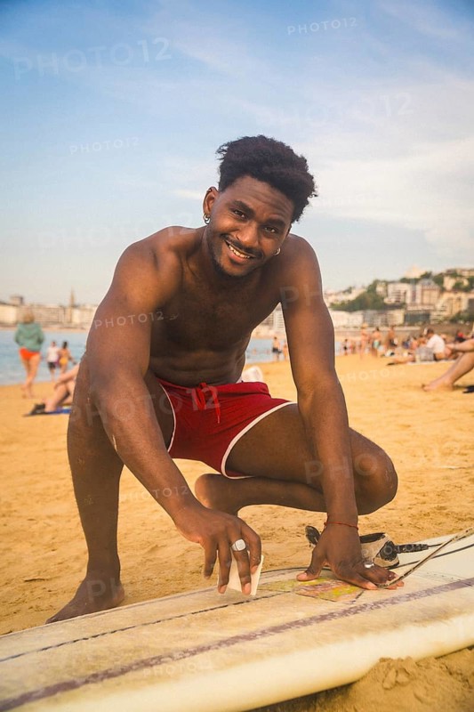 Young smiling man in red shorts applying wax to his surfboard on a sandy beach, preparing for a surf session on a warm summer day, with people and city in background