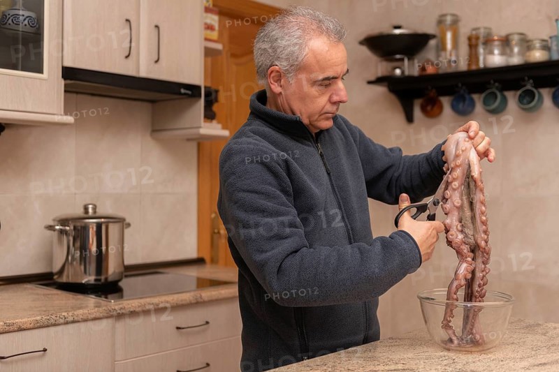 A man wearing a navy jacket is in a modern kitchen, preparing fresh octopus with scissors over a glass bowl. A stainless steel pot is visible in the background
