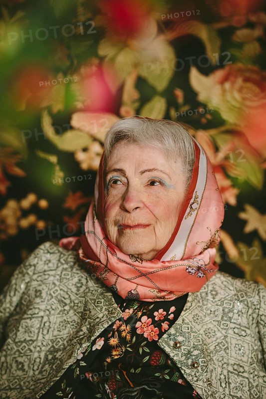 An elderly woman wearing a patterned silk scarf and jacket sits against a colorful floral backdrop. Her serene expression and the vibrant colors create a warm, studio portrait theme