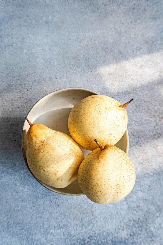 Three ripe Chinese white pears rest in a ceramic bowl placed on a textured light blue surface. The soft natural light highlights their smooth skin and subtle speckles