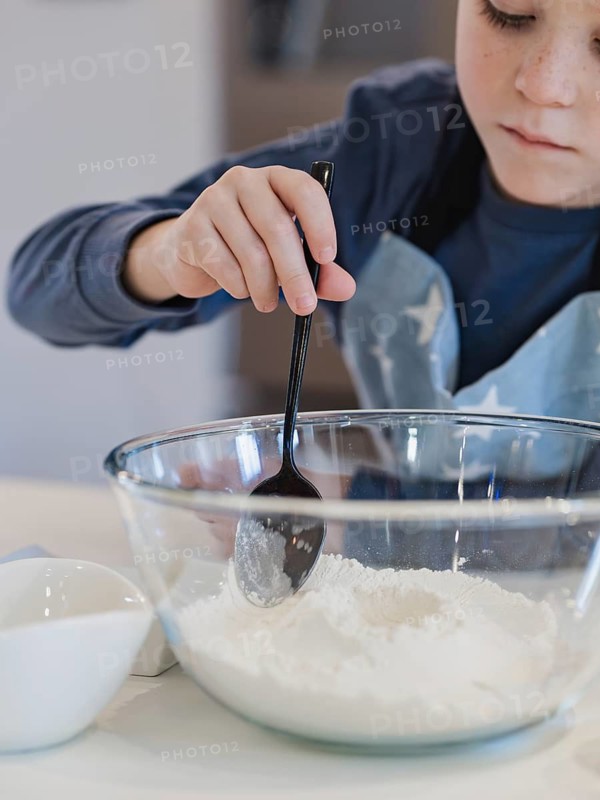 A child mixes flour in a glass bowl using a black spoon in a home kitchen. The scene captures a close up of the activity, focusing on the child hand and the baking ingredients