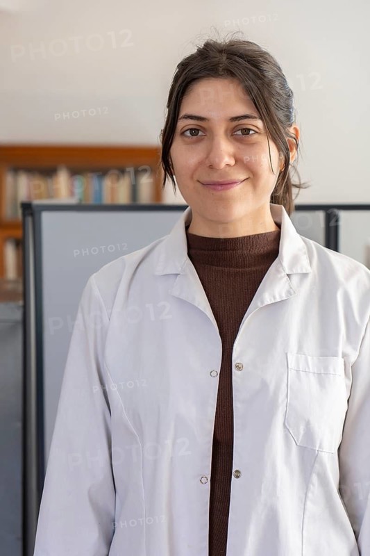 Headshot portrait of a young brunette female scientist in a white lab coat smiling gently at the camera with a blurred office bookshelf in the background
