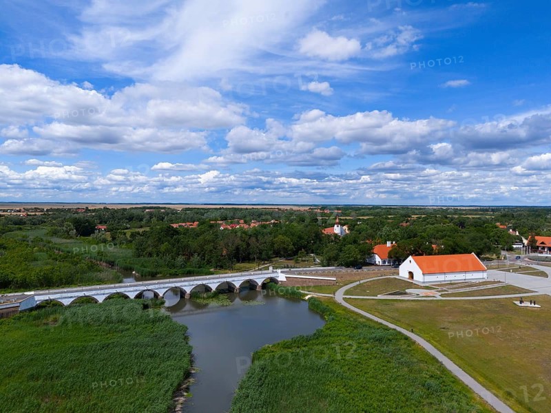 Panoramic view of a river with a bridge and a surrounding village under a wide, cloudy sky, aerial view, nine-arched stone bridge, Kilenclyukú híd, Hortobagy, Hortobagy National Park, Hortobágyi Nemzeti Park, Hortobágyi Csillagoségbolt Park, Puszta, steppe area, UNESCO Biosphere Reserve, Hungary